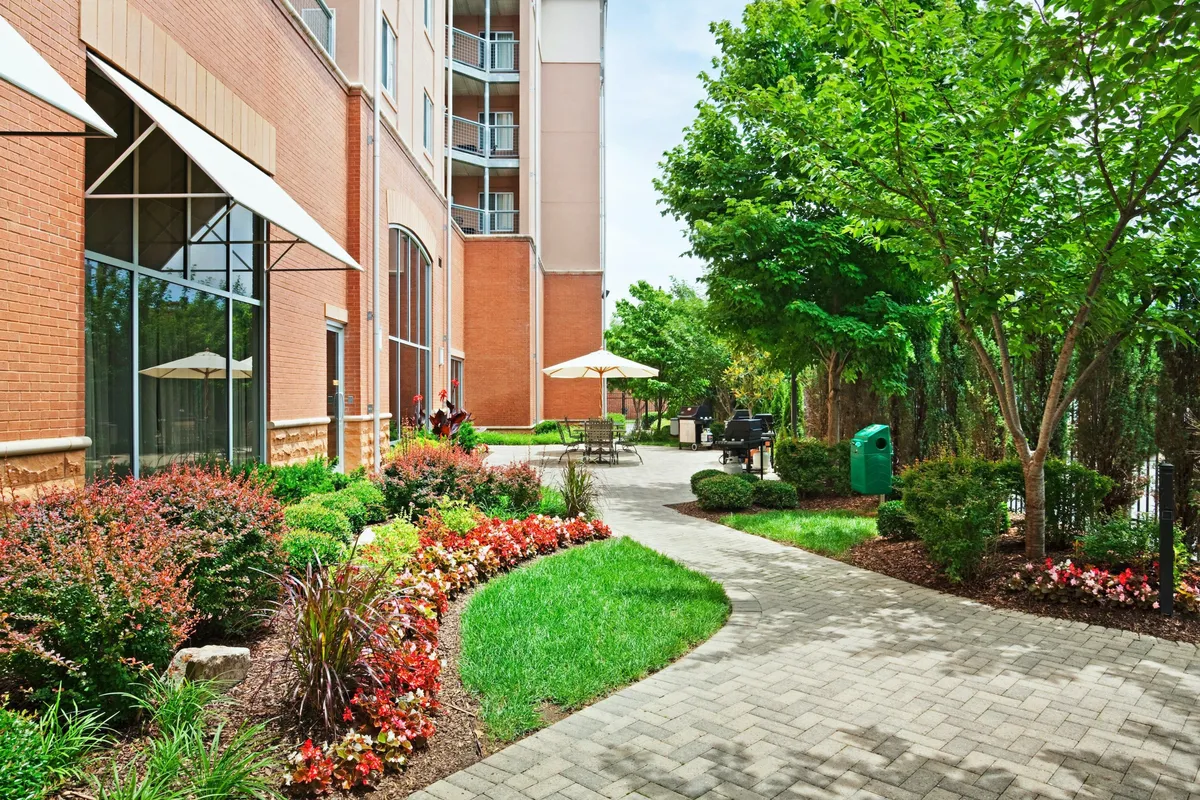 A serene outdoor area featuring lush flowerbeds, winding pathways, and inviting outdoor seating beneath green trees. The space is designed for relaxation and socializing, with a peaceful, garden-like ambiance that contrasts yet complements the urban energy of downtown.