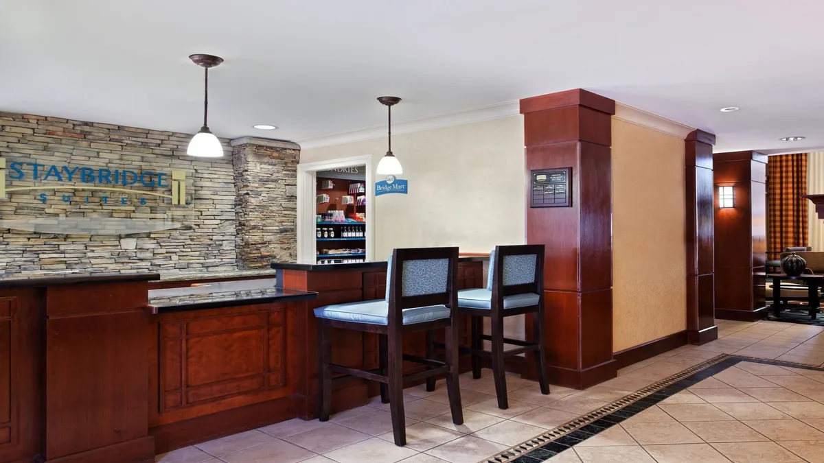 This image features the hotel's reception area with a prominent reception desk, bar stools for seating, a striking stone wall, and clear hotel signage. The space is bustling with guests checking in and others casually socializing, creating a welcoming and professional first impression.