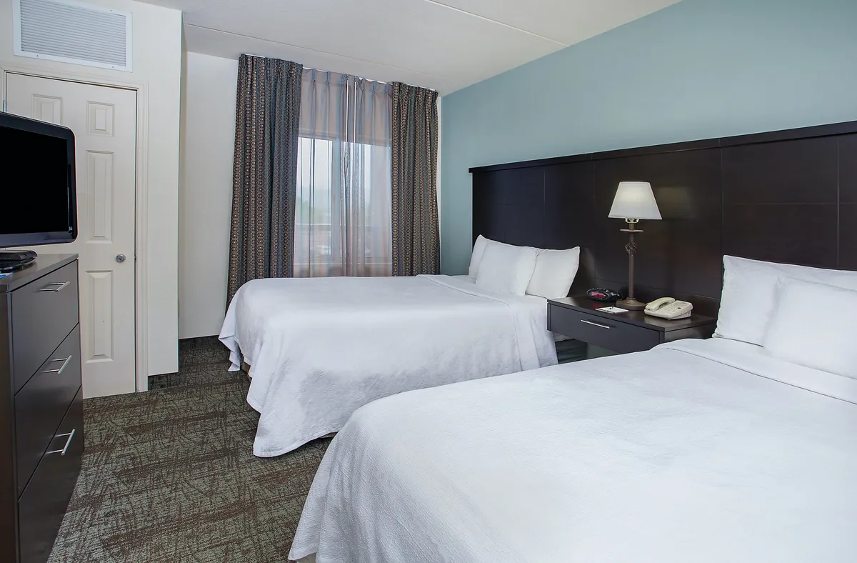 A serene bedroom scene featuring two beds, a nightstand with a lamp, a television, and curtains. The setup is relaxed, comfortable, and ideal for winding down or enjoying quiet moments.
