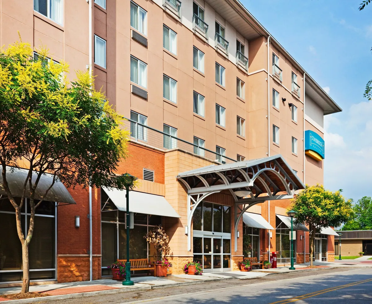 A friendly, approachable view of the building's entrance featuring an awning, abundant greenery with trees and planters, and guests socializing or relaxing outdoors. The image suggests an easy transition from the hotel to city life, with elements like outdoor seating and visible street-side access.
