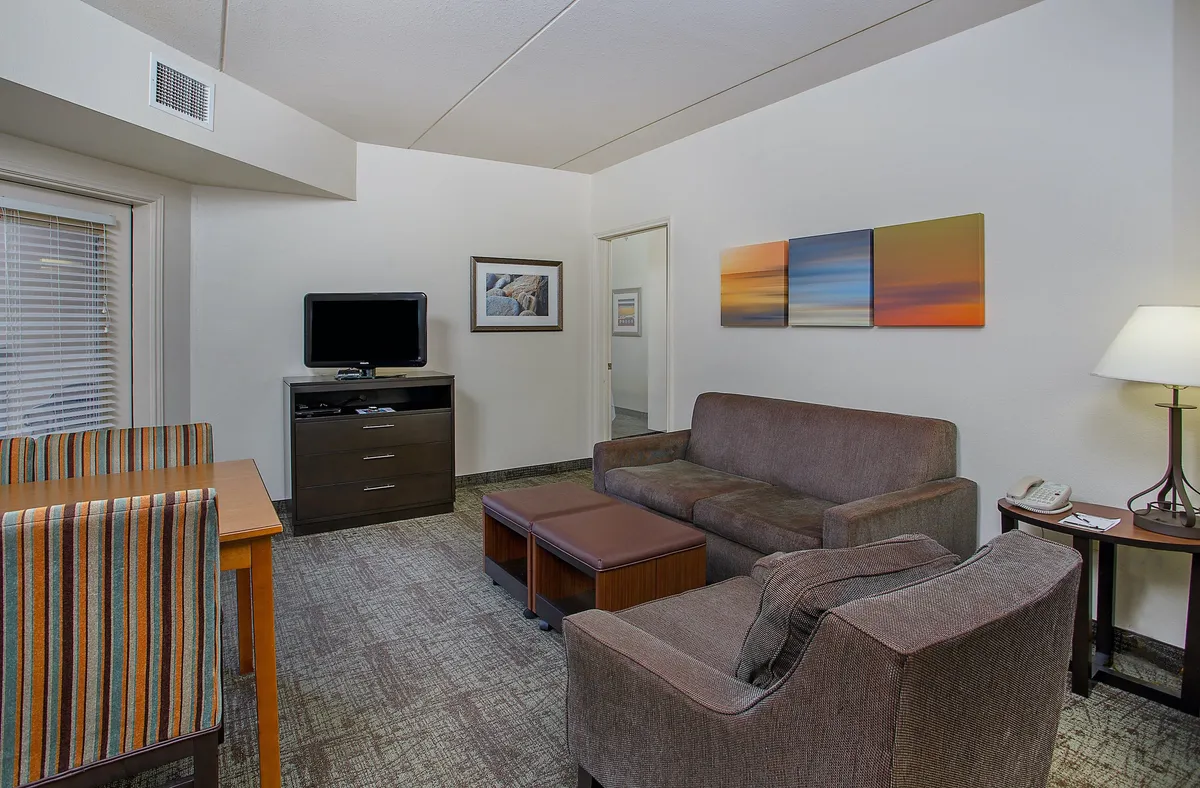 The image showcases a bright and stylish open-plan living area featuring a cozy sofa, a modern dining table set for four, a large flat-screen television, and vibrant artwork on the wall. The layout invites relaxation, conversation, and shared meals, all in a comfortable, welcoming space.
