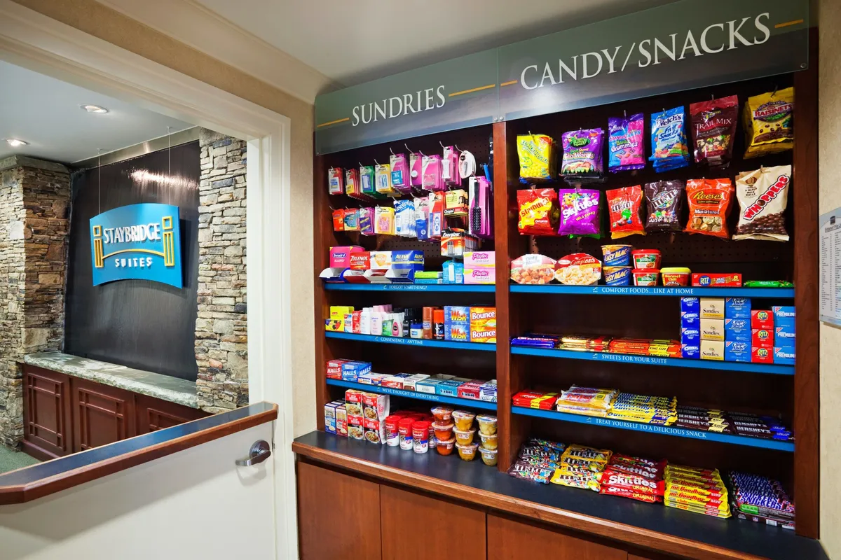 The image features a well-stocked snack and sundries shop adjacent to the Staybridge Suites sign, with shelves lined with quick bites and essentials. The wooden counter adds warmth and convenience.