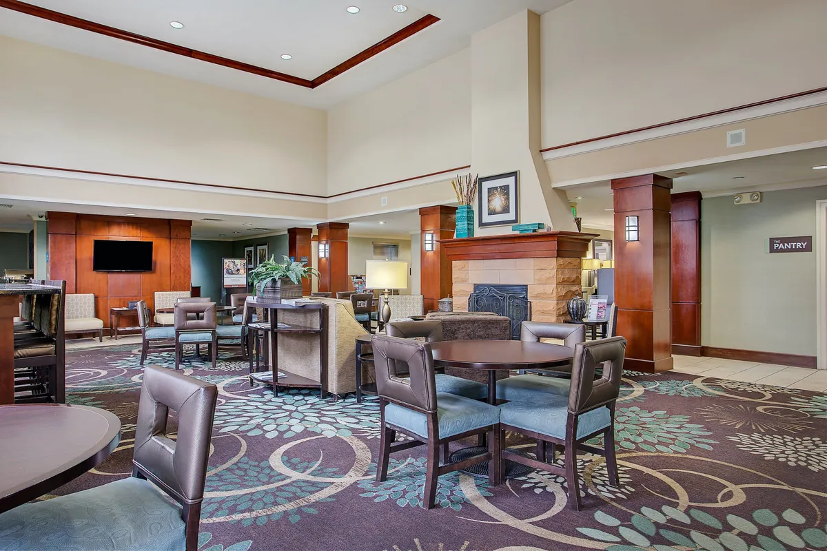 A spacious, welcoming lounge area with comfortable seating, a fireplace, decorative plants, and curated art pieces. The ambiance is warm and inviting, suggesting a place to both relax and socialize. The 'pantry' sign hints at additional amenities nearby.