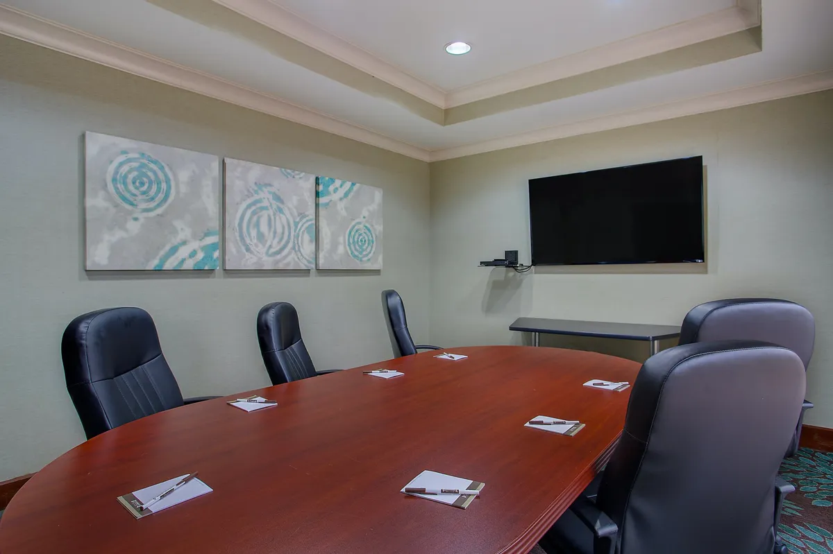 This image depicts an interior space centered around an oval wooden conference table surrounded by black leather chairs. The room also features abstract wall art and a flat-screen TV mounted on the wall, creating a professional, modern, and comfortable environment. The lighting is even, enhancing the sense of calm focus, and there are no visible exterior windows or connections to outdoor spaces.
