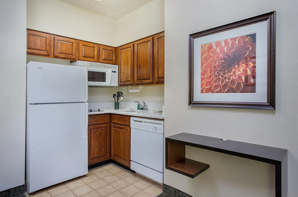 A warmly lit in-suite kitchen featuring wooden cabinets, white appliances (including a stove and microwave), and a touch of homeliness through floral artwork. The space is set up for meal preparation, cooking, and socializing, with ample counter space for culinary activities.